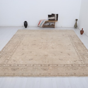 Pode incluir: Tapete bege retangular grande com um design de borda intrincado e motivos florais centrais. O tapete é colocado em um chão branco, com itens decorativos no fundo, sugerindo um ambiente de sala de estar.