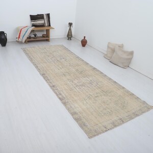 May include: A rectangular, beige runner rug with a faded, patterned design. The rug has a dark border and is placed on a white wooden floor. Decorative items and pillows are in the background.