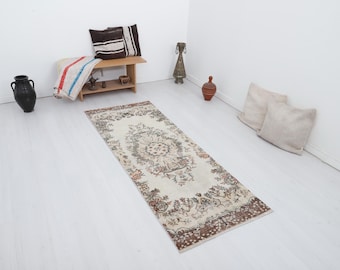 May include: A rectangular, cream-colored rug with a detailed floral pattern in shades of brown, beige, and peach. The rug is placed on a white floor, with other decorative items in the background, including pillows and a bench.