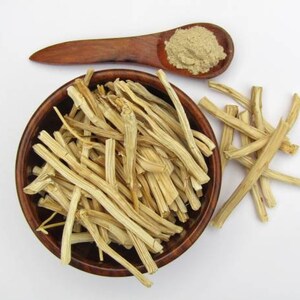 May include: A wooden bowl filled with dried, light brown, fibrous plant stems. A wooden spoon with a light brown powder is next to the bowl. A few stems are scattered on the white surface.