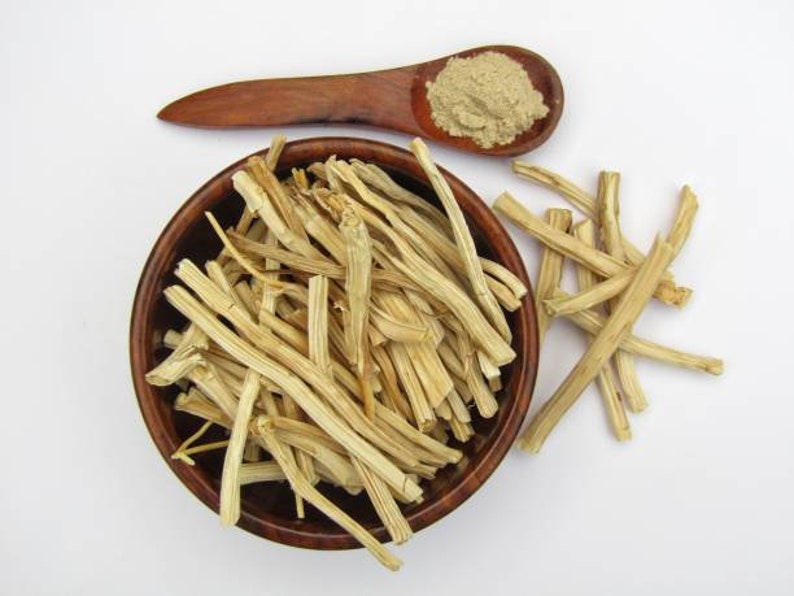 May include: A wooden bowl filled with dried, light brown, fibrous plant stems. A wooden spoon with a light brown powder is next to the bowl. A few stems are scattered on the white surface.