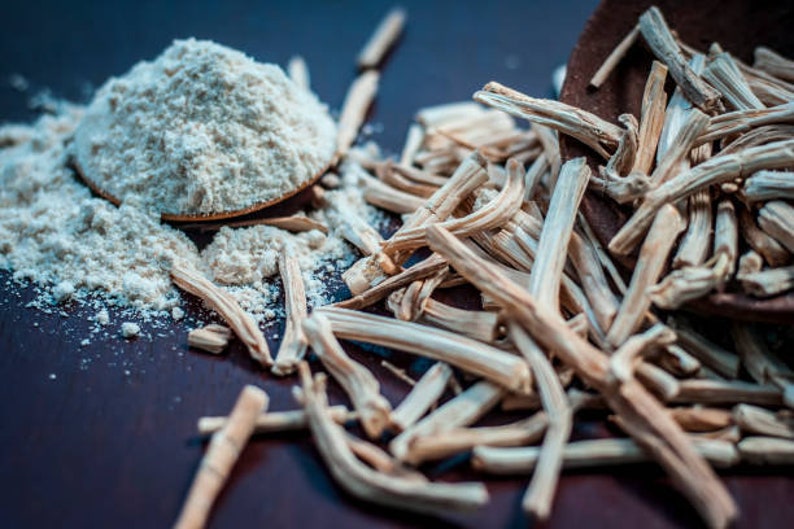 May include: A wooden spoon filled with white powder, surrounded by dried, light brown sticks on a dark brown surface.