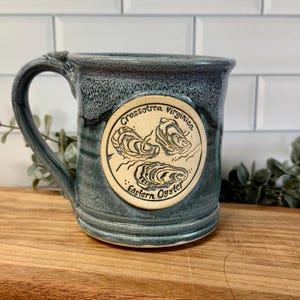 May include: A ceramic mug with a blue-grey mottled glaze featuring a circular cream-colored medallion on the front. The medallion displays a black line drawing of three oysters with the text Crassostrea Virginica Eastern Oyster printed around them.