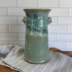 May include: A handcrafted ceramic vase in shades of teal and green. The vase has a cylindrical shape with two small handles and a slightly flared rim. The glaze has a mottled appearance, adding to its unique, artisanal charm.