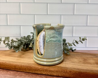 May include: A cylindrical ceramic sponge holder with a glossy, variegated blue-green glaze and a wide vertical cutout on the front. The holder features a raised base rim and contains a round, textured, light-colored cleaning sponge inside.