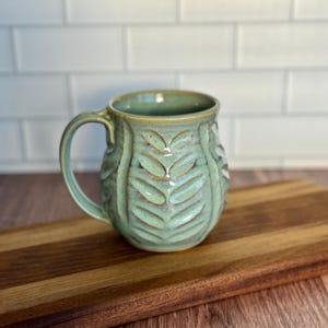 May include: A light green ceramic mug with a leaf design. The mug has a rounded body and a handle.