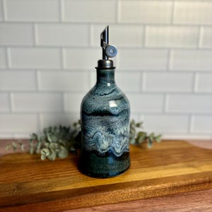 May include: A ceramic oil bottle with a stainless steel spout. The bottle has a dark blue and teal glaze with a layered effect. The bottle is on a wooden cutting board.