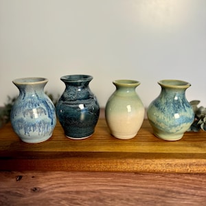 May include: Five small ceramic vases in various shades of blue and green with white and brown accents. The vases are arranged in a row on a wooden surface.