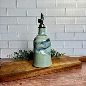 May include: A ceramic olive oil bottle with a blue and green glaze. The bottle has a metal pour spout and is sitting on a wooden cutting board.