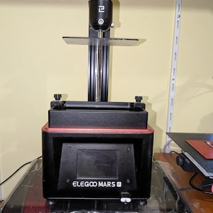 May include: A black ELEGOO MARS P 3D printer with a red trim. The printer has a clear platform, a black adjustable arm, and a small screen displaying information. The printer is on a table.