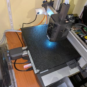 Peut inclure: Une imprimante 3D avec un plateau d'impression noir et une tête d'impression gris foncé. L'imprimante dispose d'un écran tactile et est connectée à divers câbles. La marque "ANYCUBIC" est visible sur le côté.