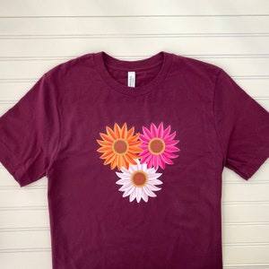 May include: A maroon t-shirt with a design of three flowers. The flowers are orange, pink, and white.