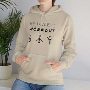 Könnte beinhalten: Ein beiger Hoodie mit dem Text "My Favorite Workout" und drei schwarzen Silhouetten eines Korkenziehers in verschiedenen Positionen.