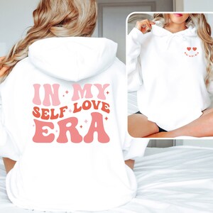 Könnte beinhalten: Weißer Hoodie mit dem Text "In My Self Love Era" in rosa und roten Buchstaben auf der Rückseite. Die Vorderseite des Hoodies hat zwei kleine rosa Herzen mit dem Text "Self Love Era" in kleineren Buchstaben.
