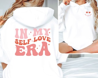 May include: White hoodie with the text "In My Self Love Era" in pink and red lettering on the back. The front of the hoodie has two small pink hearts with the text "Self Love Era" in smaller lettering.