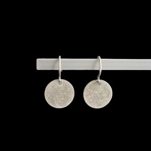 Peut inclure: Une paire de boucles d'oreilles en argent avec un motif circulaire texturé. Les boucles d'oreilles sont suspendues à une barre blanche.