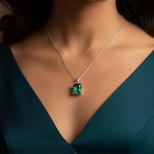 Puede incluir: Un collar de plata con un colgante rectangular verde esmeralda. El colgante está engastado en un marco de plata y cuelga de una fina cadena de plata. El collar se muestra sobre un fondo verde azulado oscuro.