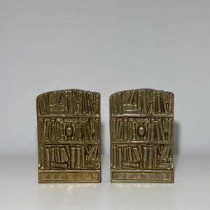 Puede incluir: Dos sujetalibros de metal dorado, cada uno dise&ntilde;ado para parecerse a una estanter&iacute;a. Los sujetalibros son rectangulares con una parte superior curvada y presentan representaciones detalladas de libros y un jarr&oacute;n decorativo. Los sujetalibros est&aacute;n colocados sobre una superficie blanca.