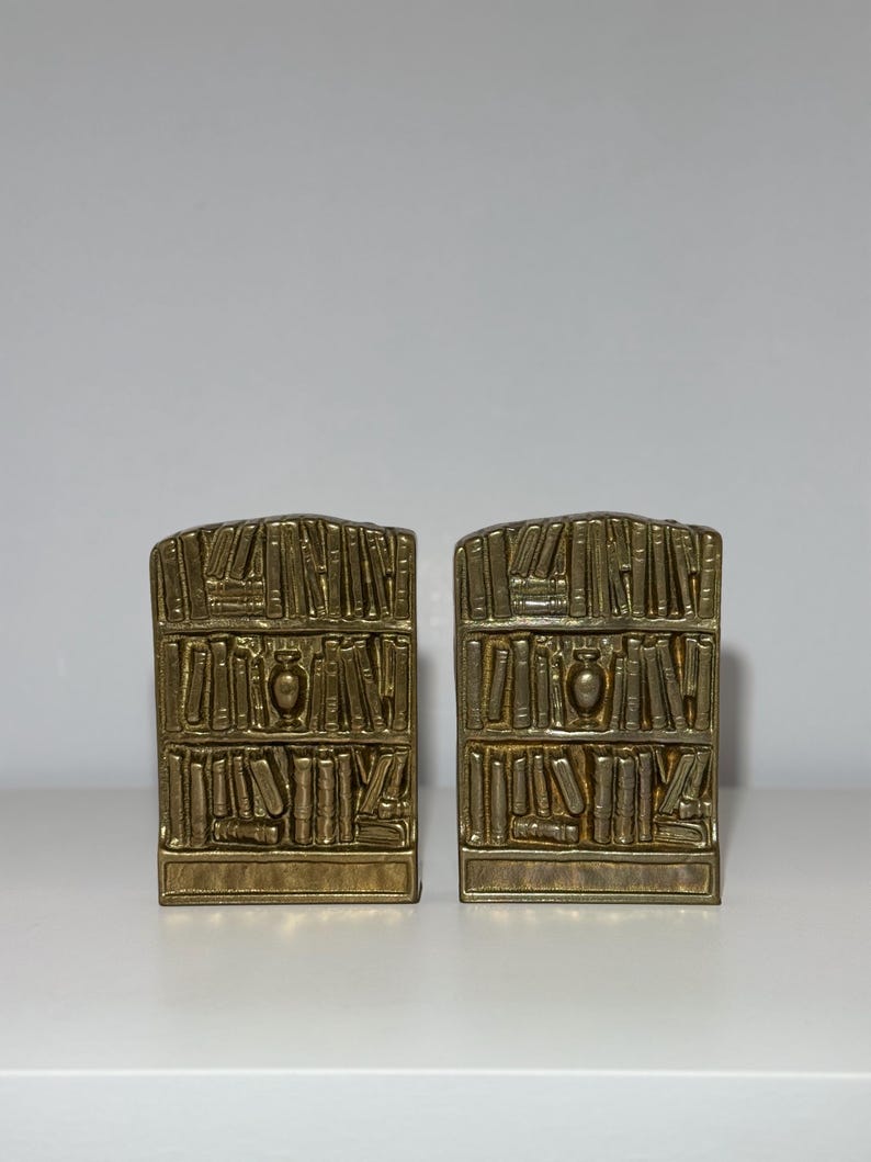 Puede incluir: Dos sujetalibros de metal dorado, cada uno dise&ntilde;ado para parecerse a una estanter&iacute;a. Los sujetalibros son rectangulares con una parte superior curvada y presentan representaciones detalladas de libros y un jarr&oacute;n decorativo. Los sujetalibros est&aacute;n colocados sobre una superficie blanca.