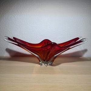 Könnte beinhalten: Eine leuchtend rote Glasschale mit einem einzigartigen, skulpturalen Design. Die Schale hat eine ausgestellte, blütenblattartige Form mit einem klaren Glassockel. Die Farbe geht von einem tiefen Rot in einen helleren Orangeton zur Mitte über. Die Schale steht auf einer hellen Holzoberfläche.