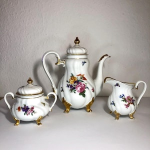 May include: A white porcelain tea set with gold trim and floral designs. The set includes a teapot, a sugar bowl with a lid, and a creamer. The teapot and sugar bowl have gold finials. The set is on a white surface.