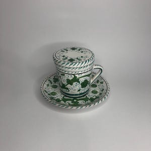 May include: A white ceramic teacup and saucer with a green floral design. The cup has a lid.