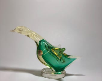 Puede incluir: Una figura decorativa de pato de cristal. El pato presenta una cabeza y un pico transparentes, un cuerpo verde con detalles amarillos y una base transparente. La escultura de cristal tiene una base circular y un pequeño acento redondo de color dorado en su costado.