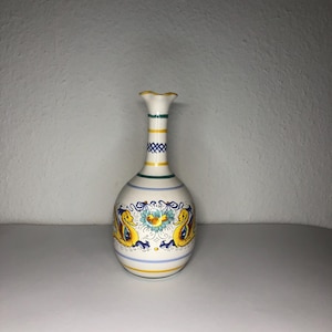 May include: A white ceramic vase with a yellow, blue and green floral design. The vase has a narrow neck and a rounded body.
