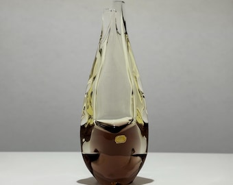 May include: A clear glass vase in a teardrop shape. The vase has a brown base that fades to a yellow-tinted top. The vase is on a white surface.