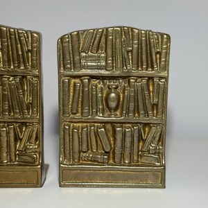Puede incluir: Un par de sujetalibros dorados, cada uno con la forma de una estanter&iacute;a llena de libros. Los sujetalibros tienen una base rectangular y una parte superior curvada, con un peque&ntilde;o jarr&oacute;n en el centro del dise&ntilde;o de la estanter&iacute;a.