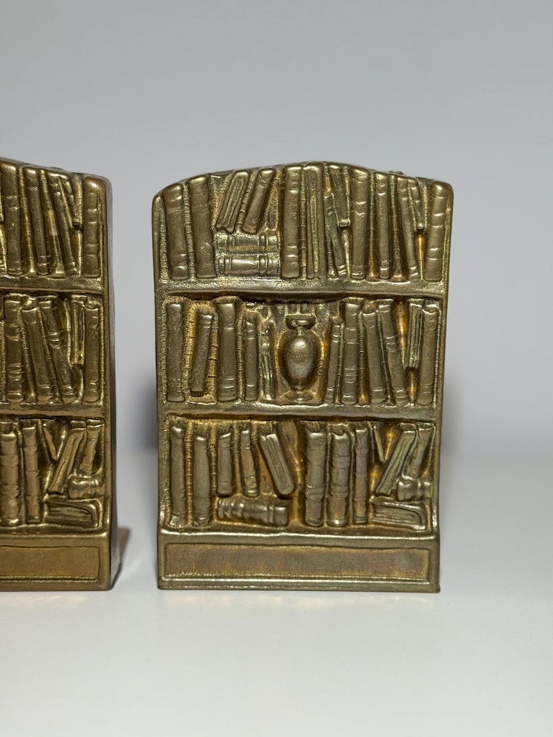 Puede incluir: Un par de sujetalibros dorados, cada uno con la forma de una estanter&iacute;a llena de libros. Los sujetalibros tienen una base rectangular y una parte superior curvada, con un peque&ntilde;o jarr&oacute;n en el centro del dise&ntilde;o de la estanter&iacute;a.