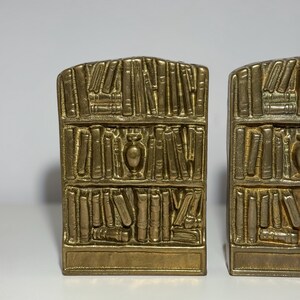 Puede incluir: Dos sujetalibros dorados, cada uno dise&ntilde;ado para parecerse a una estanter&iacute;a llena de libros. Los sujetalibros tienen una base rectangular y una parte superior curvada, con representaciones detalladas de libros y un jarr&oacute;n decorativo. El dise&ntilde;o es vintage y decorativo.
