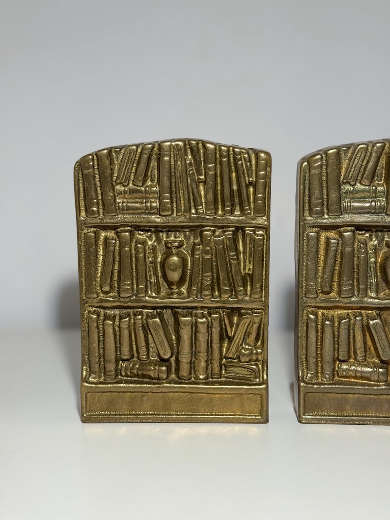 Puede incluir: Dos sujetalibros dorados, cada uno dise&ntilde;ado para parecerse a una estanter&iacute;a llena de libros. Los sujetalibros tienen una base rectangular y una parte superior curvada, con representaciones detalladas de libros y un jarr&oacute;n decorativo. El dise&ntilde;o es vintage y decorativo.