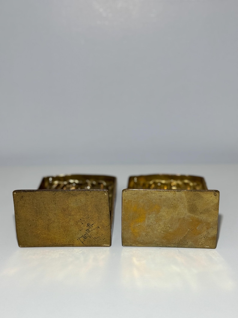 Puede incluir: Cuatro sujetalibros de lat&oacute;n antiguos. Cada sujetalibros tiene una base rectangular y una parte superior decorativa con un patr&oacute;n texturizado. El lat&oacute;n tiene una p&aacute;tina c&aacute;lida y envejecida. Los sujetalibros se exhiben sobre una superficie blanca.