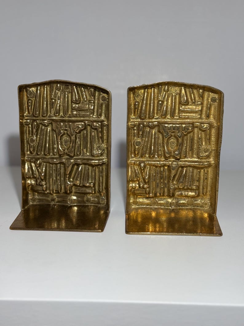 Puede incluir: Un par de sujetalibros de metal color dorado. Cada sujetalibros tiene forma rectangular con un dise&ntilde;o en relieve detallado que se asemeja a una pila de libros. Los sujetalibros est&aacute;n dise&ntilde;ados para sostener libros en un estante.