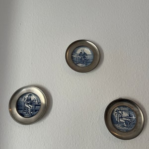 Könnte beinhalten: Drei dekorative Teller mit silbernen Rändern und blauen und weißen Designs, die Menschen zeigen, die an Tischen sitzen.