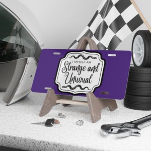 May include: Purple license plate with a white border and a black and white graphic that says "I Myself Am Strange and Unusual" with a snake design. The license plate is on a wooden stand.