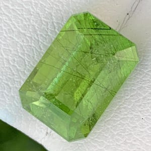 May include: A rectangular-cut gemstone in a vivid, translucent green. The gem features fine, dark inclusions, creating a distinctive internal pattern. The facets of the cut enhance its brilliance and clarity. Suitable for jewellery.
