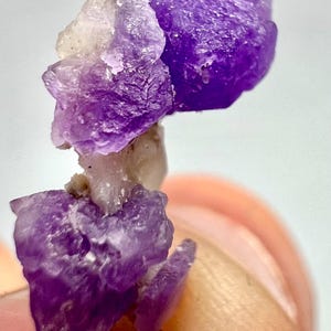 May include: Close-up of a cluster of raw amethyst crystals. The crystals display a deep purple hue with some lighter, translucent areas. The rough texture and natural formation of the amethyst are clearly visible, showcasing its geological characteristics.