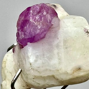 May include: A close-up of a natural mineral specimen featuring a vibrant, crystalline, magenta-colored ruby atop a creamy white matrix. The ruby's facets catch the light, contrasting with the opaque base. The specimen is mounted on a metal stand.