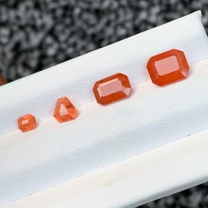 以下が含まれることがあります： 白地の表面に、さまざまな形とサイズの4つのオレンジ色の宝石が表示されています。宝石には、正方形、三角形、長方形、エメラルドカットが含まれます。宝石は鮮やかなオレンジ色です。