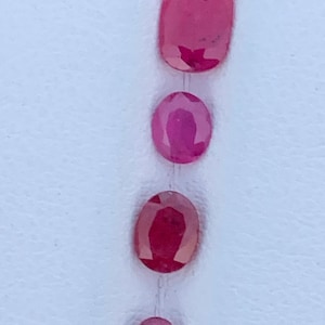 May include: Seven oval-shaped pink gemstones of varying sizes arranged vertically on a white surface.