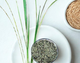 May include: A white plate with a small glass bowl of dried herbs, with green grass blades. A small bowl of brown twine is in the background. The image is set against a white background. The herbs and twine are likely for cooking.