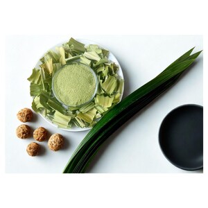 Peut inclure: Vue de dessus sur une surface blanche avec une assiette de feuilles et de poudre de pandan vertes, une longue feuille verte, une assiette noire et plusieurs objets ronds bruns. La poudre est dans un bol transparent.