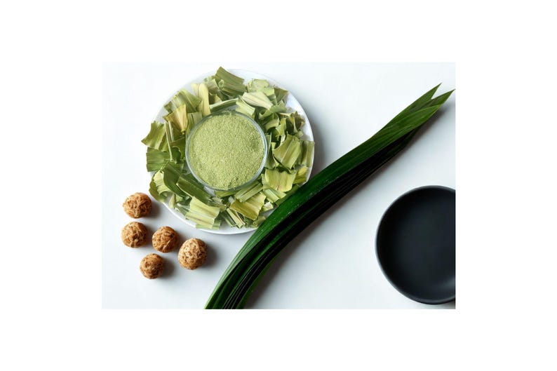 Peut inclure: Vue de dessus sur une surface blanche avec une assiette de feuilles et de poudre de pandan vertes, une longue feuille verte, une assiette noire et plusieurs objets ronds bruns. La poudre est dans un bol transparent.