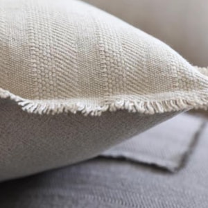 Pode incluir: Uma almofada decorativa bege com tecido texturizado, um padrão subtil de espinha de peixe e bordas desfiadas. A almofada tem uma aparência suave e natural com linhas de tecelagem verticais visíveis e um detalhe de franjas na costura.