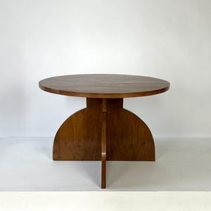 May include: A round wooden table with a smooth, circular top and a base constructed from two intersecting vertical panels that form a cross shape, featuring a warm, medium-brown wood grain finish throughout the entire structure.