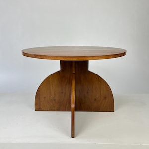 May include: A round wooden table with a smooth, circular top and a base constructed from two intersecting vertical panels that form a cross-shaped support structure. The wood features a warm, medium-brown tone with visible grain patterns.
