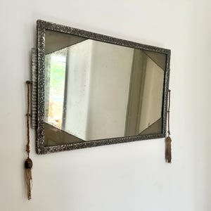Peut inclure: Miroir rectangulaire avec un cadre argenté texturé et des accents géométriques. Le miroir est accroché à un mur blanc et comporte des glands décoratifs de chaque côté. Le design est vintage et ajoute une touche d'élégance.