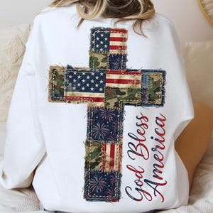 Puede incluir: Sudadera blanca con un diseño de cruz en la espalda, compuesta por parches de la bandera estadounidense y tela de camuflaje. Las palabras "God Bless America" están impresas en escritura roja y azul. La sudadera es de estilo casual.
